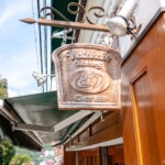 A wooden sign shaped like a bread slice hangs outside Hakone Watanabe Bakery, reading Watanabe Bakery Since 1897, with a loaf icon carved in the center. The brown and white building sits among trees in the background.