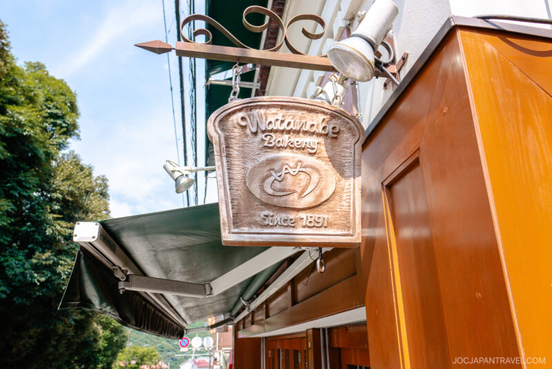 A wooden sign shaped like a bread slice hangs outside Hakone Watanabe Bakery, reading Watanabe Bakery Since 1897, with a loaf icon carved in the center. The brown and white building sits among trees in the background.