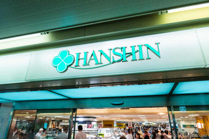 A store entrance with a sign reading HANSHIN in teal letters and a clover logo above, welcoming shoppers into the brightly lit interior—a vibrant scene at the Hankyu and Hanshin Department Stores Osaka.