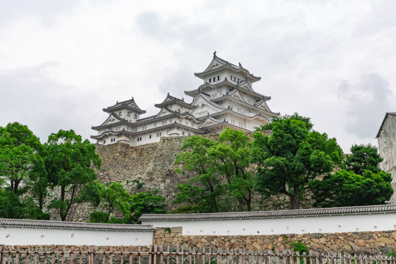 Himeji Castle is a white traditional Japanese castle with multiple roofs, standing on a stone foundation, surrounded by green trees and a stone wall under a cloudy sky.