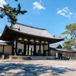 Nara Horyu-ji’s traditional Japanese temple gate features a tiled roof, wooden pillars, and stone steps, nestled among pine trees beneath a bright blue sky with scattered clouds.