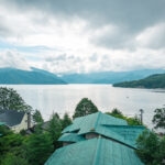 Hoshino Resorts KAI Nikko (星野リゾート 界 日光) – A Luxury Ryokan by Lake Chuzenji A peaceful lakeside scene with green-roofed buildings, including Hoshino Resorts KAI Nikko in the foreground, surrounded by lush trees and distant mountains under a partly cloudy sky reflected in the calm lake.