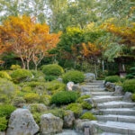 A stone staircase winds through a lush Japanese garden at Hotel Kaneyamaen Yamanashi, with neatly trimmed bushes, rocks, and trees with green and red foliage, all surrounded by tall trees in the background.
