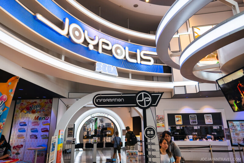 Entrance to Joypolis DECKS, a SEGA indoor amusement park, featuring a large neon sign overhead, an information booth, ticket counter, and several visitors exploring the modern, colorful interior.