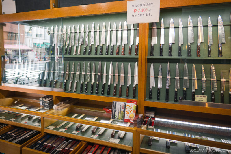 A glass display case in Kappabashi Kamata Knife Shop showcases a wide variety of Japanese kitchen knives, neatly arranged in rows, with boxed knives and sharpeners on the lower shelves and a sign in Japanese above.