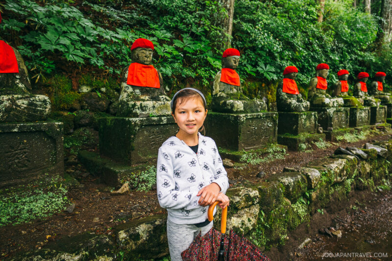 A smiling child stands in front of a row of stone statues wearing red hats and bibs at Nikko Kanmangafuchi Abyss. In the lush, green setting, the child holds an umbrella and wears a light hoodie with a skull pattern.