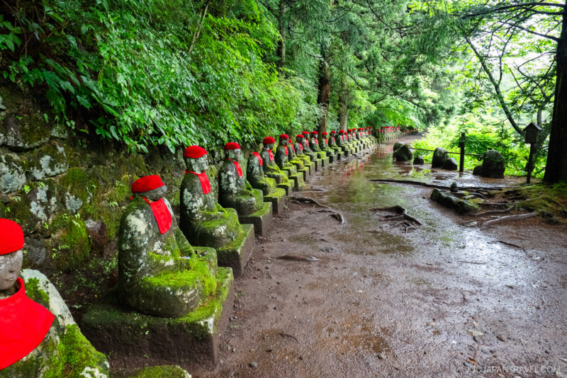 A long row of moss-covered stone statues wearing red hats and bibs lines a forest path at Nikko Kanmangafuchi Abyss, surrounded by lush green trees and wet ground.