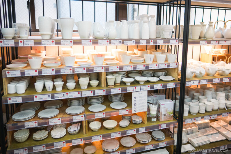 Shelves filled with neatly arranged white ceramic dishes, bowls, cups, and plates of various shapes and sizes in a well-lit Kappabashi Baise store.