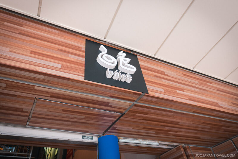 A sign with stylized white Japanese characters and the words Kappabashi Baise below them is mounted on a wood-paneled exterior wall near a ceiling.