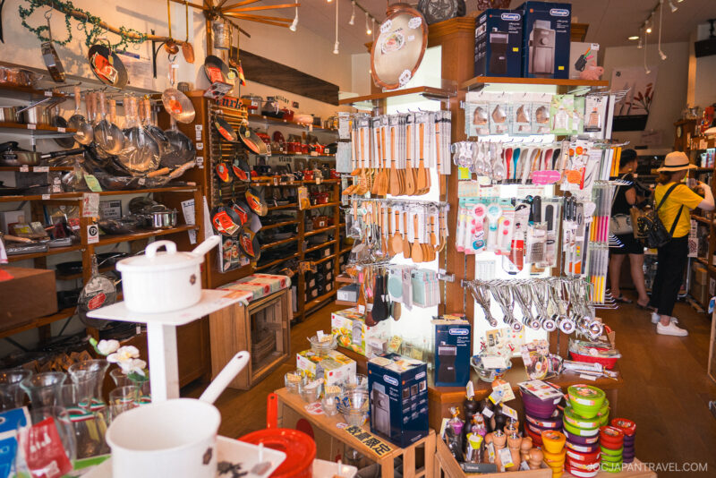 A brightly lit shop, Kappabashi Dr. Goods, filled with kitchenware, utensils, pots, pans, and various cooking tools displayed on shelves and racks. Two shoppers are browsing in the background.