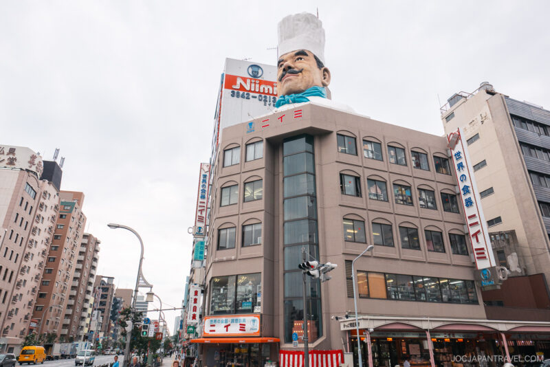 A multi-story building in Tokyo’s Kappabashi district features a giant chef’s head wearing a white hat on its roof. The facade displays the Niimi sign and is surrounded by other urban buildings under a cloudy sky.