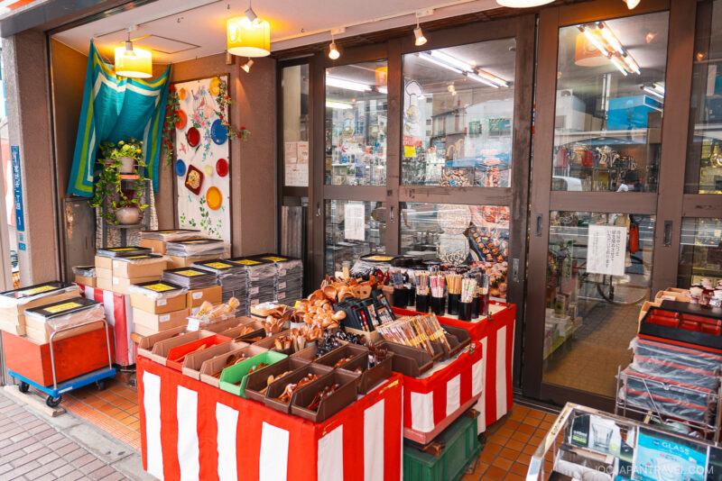 A small storefront near Kappabashi Niimi displays boxes of brushes, stationery, and various goods on tables with red-and-white striped coverings. The glass doors reveal more items inside, with bright lighting and colorful decor on the wall.