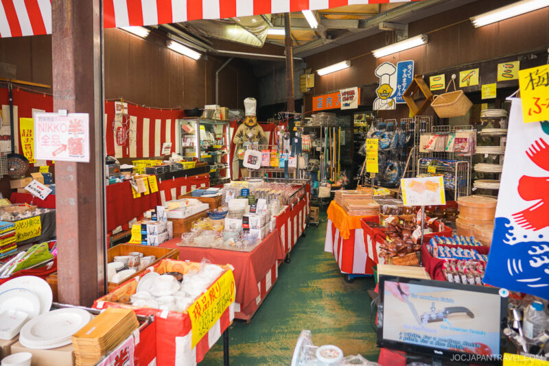 A colorful Japanese market stall near Kappabashi Niimi displays dishes, kitchenware, packaged food, and signs with Japanese writing. Red and white striped decorations hang above shelves and tables filled with various goods for sale.