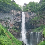 Kegon Falls (華厳滝) – Nikko’s Most Famous Waterfall Nikko Kegon Falls cascades down a rocky cliff surrounded by lush green trees and vegetation, with mist rising at the base. The vibrant scene evokes a sense of tranquility and grandeur in this breathtaking natural wonder.