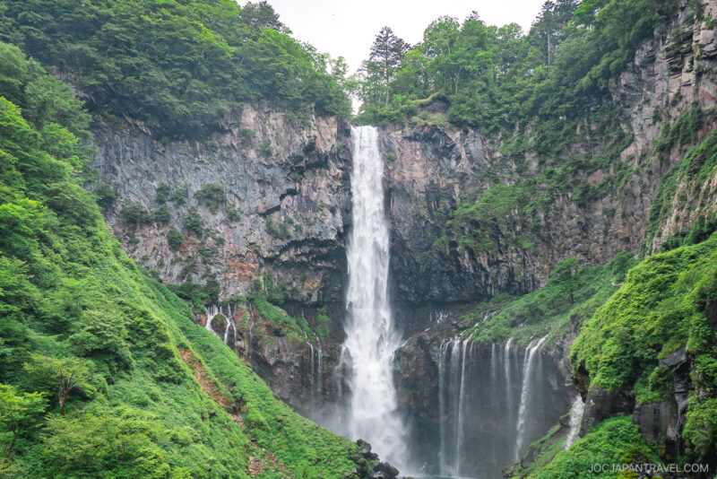 Nikko Kegon Falls cascades down a rocky cliff surrounded by lush green trees and vegetation, with mist rising at the base. The vibrant scene evokes a sense of tranquility and grandeur in this breathtaking natural wonder.