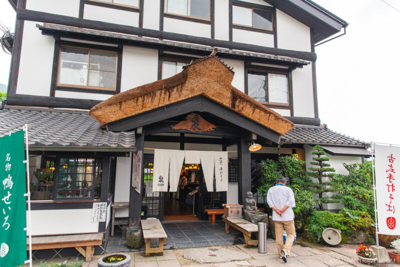 A man walks toward the entrance of a traditional Japanese building near Kirin Lake, with white walls, dark wooden beams, a thatched awning, and hanging white noren curtains. Lush greenery and banners decorate the exterior.