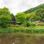 A scenic view of Kirin Lake, surrounded by lush greenery, with a café and outdoor seating on the left and a thatched-roof house in the background, set against forested hills under a cloudy sky.