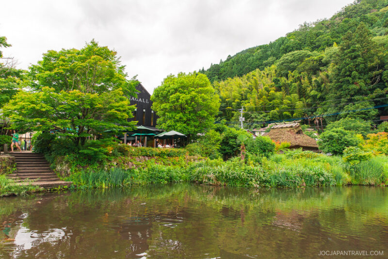 A scenic view of Kirin Lake, surrounded by lush greenery, with a café and outdoor seating on the left and a thatched-roof house in the background, set against forested hills under a cloudy sky.