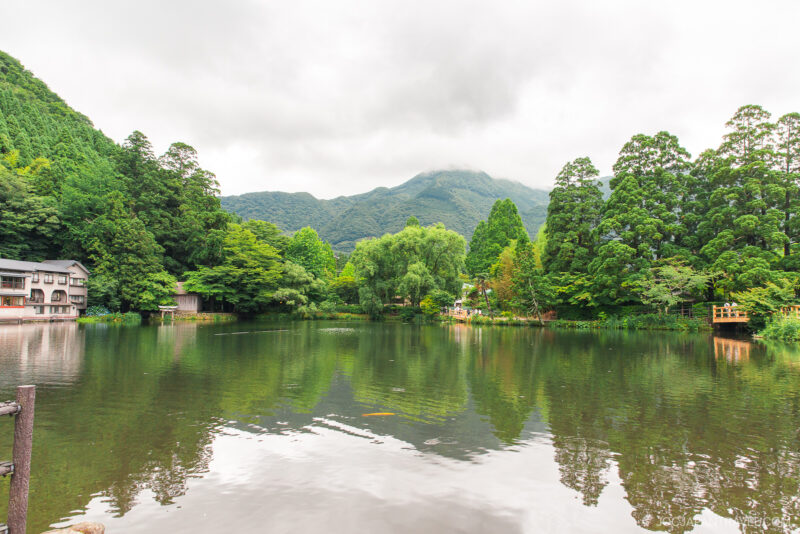 A calm Kirin Lake reflects lush green trees and mountains under a cloudy sky, with traditional buildings and a small dock at the water’s edge.