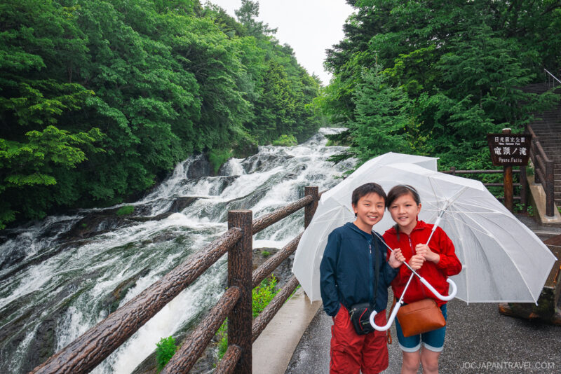 Two children stand together under a transparent umbrella on a wet path beside a wooden railing, with Lake Chuzenji’s lush green forest and a cascading waterfall in the background.