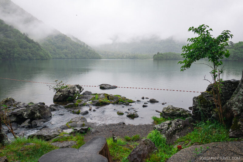 A misty Lake Chuzenji is surrounded by green forested hills, with rocks and a small tree in the foreground. A thin red-and-white rope stretches across the water, under an overcast sky with low-hanging clouds.
