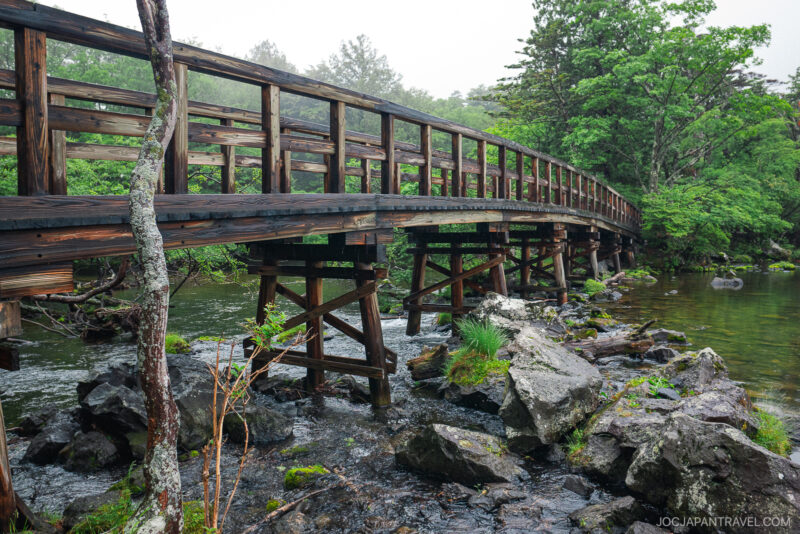 A wooden bridge crosses over a shallow, rocky stream near Lake Chuzenji, surrounded by lush green trees and foliage on a misty day.
