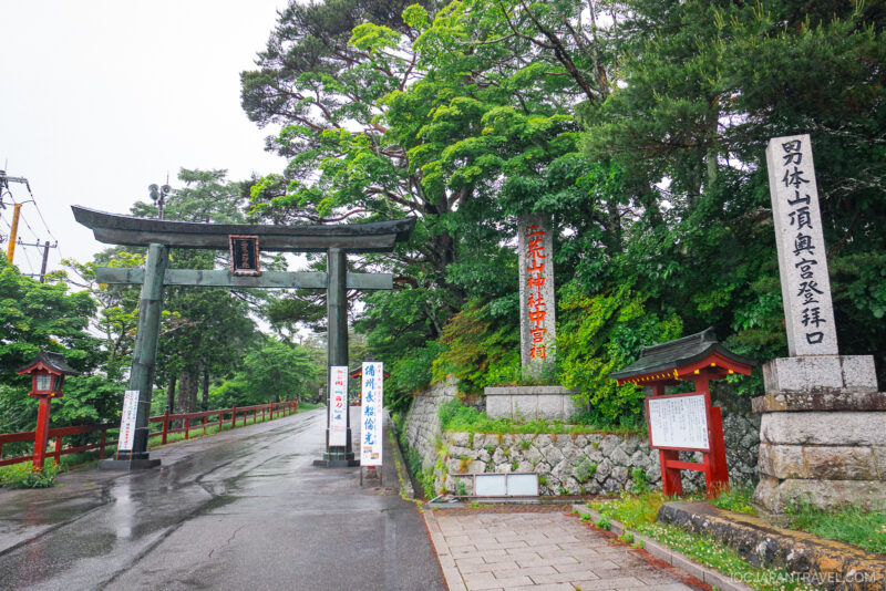A large traditional Japanese torii gate stands at the entrance to a scenic, tree-lined path near Lake Chuzenji, with stone lanterns and multiple white signs with red and black Japanese writing. The atmosphere is serene and slightly misty.