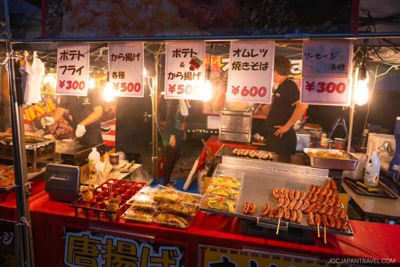 A Japanese street food stall at night, near the Lake Kawaguchi Fireworks Festival, displays fried foods and skewers. Bright signs list menu items and prices in yen above the counter as people cook behind the vibrant stall.