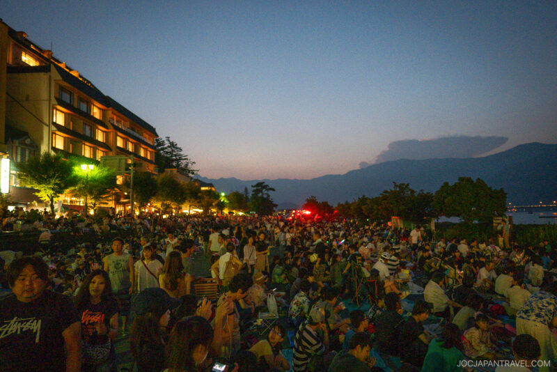 A large crowd gathers outdoors at dusk near a lit-up building, with mountains and a body of water visible in the background. The scene captures the vibrant atmosphere of the Lake Kawaguchi Fireworks Festival as evening approaches.