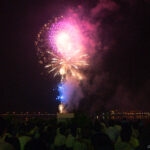 Lake Kawaguchi Fireworks Festival (河口湖湖上祭) – Fireworks with Mt. Fuji Views Colorful fireworks light up the night sky above a river at the Lake Kawaguchi Fireworks Festival, with a large crowd watching the display. City lights shimmer in the background, reflecting beautifully on the water.