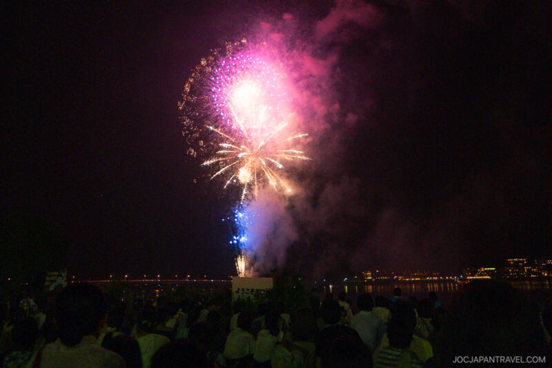 Colorful fireworks light up the night sky above a river at the Lake Kawaguchi Fireworks Festival, with a large crowd watching the display. City lights shimmer in the background, reflecting beautifully on the water.