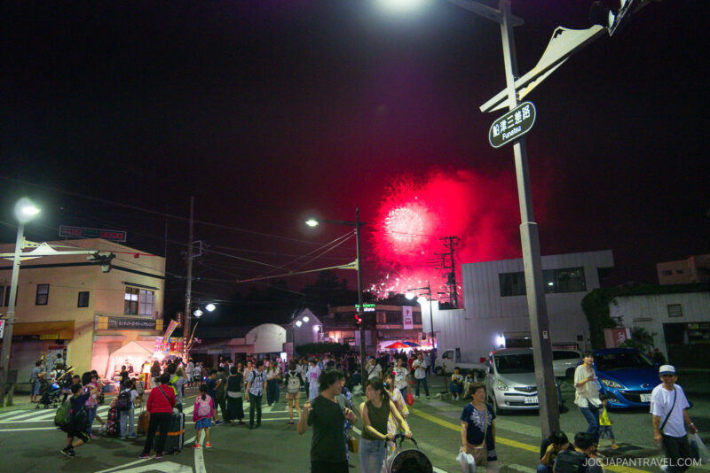 A lively street scene at night with crowds of people watching red fireworks from the Lake Kawaguchi Fireworks Festival light up the sky. Buildings, cars, and streetlights add to the festive urban atmosphere.