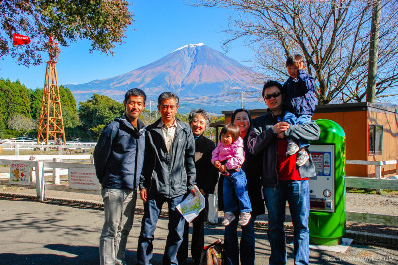 A group of seven people, including children, pose and smile outdoors at Makaino Farm Resort Shizuoka with Mount Fuji in the background on a sunny day. Trees with autumn leaves and a green vending machine are nearby.