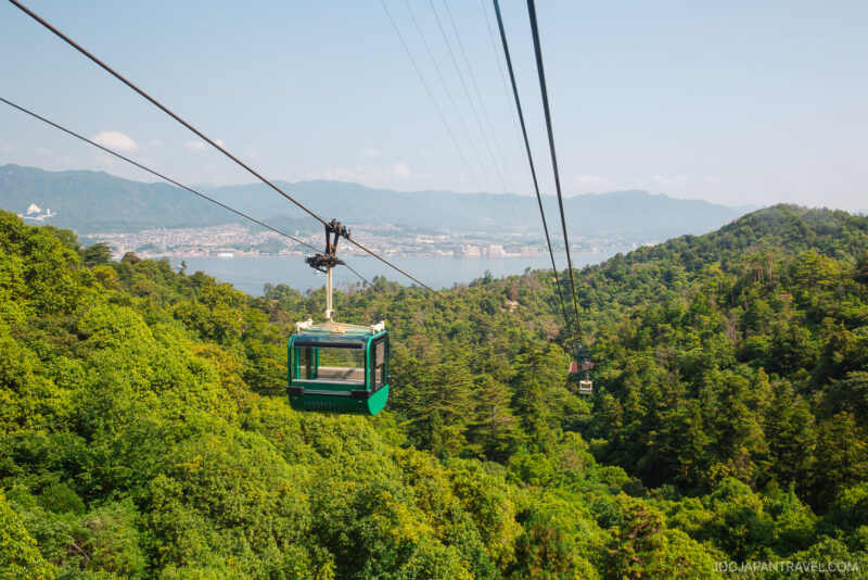 A green cable car travels above the lush forested hills of Mount Misen, with a distant city and mountains visible across a body of water under a clear blue sky.