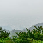 Exploring Mt. Rokko (六甲山) – Scenic Ropeway Adventure from Arima Onsen A hazy cityscape is partially obscured by mist, with lush green trees and forested hills of Mount Rokko in the foreground. The city buildings are barely visible through the fog.