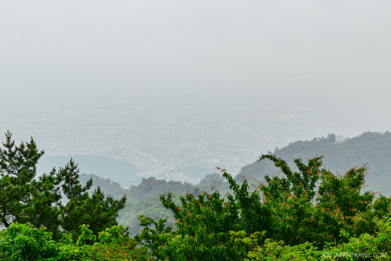 A hazy cityscape is partially obscured by mist, with lush green trees and forested hills of Mount Rokko in the foreground. The city buildings are barely visible through the fog.