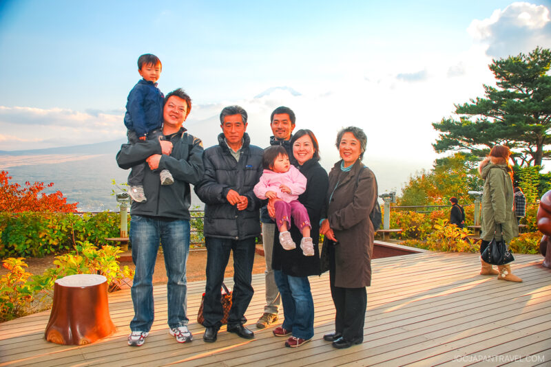 A group of seven people, including adults and children, pose and smile on a scenic wooden deck surrounded by trees and autumn foliage near the Mt. Fuji Panoramic Ropeway, with a hazy landscape visible in the background.