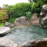 An outdoor Japanese onsen at Musouen Hotel, surrounded by large rocks, green bushes, and a bamboo fence. Steam rises gently from the clear hot spring water, with a wooden bucket resting at the pools edge.