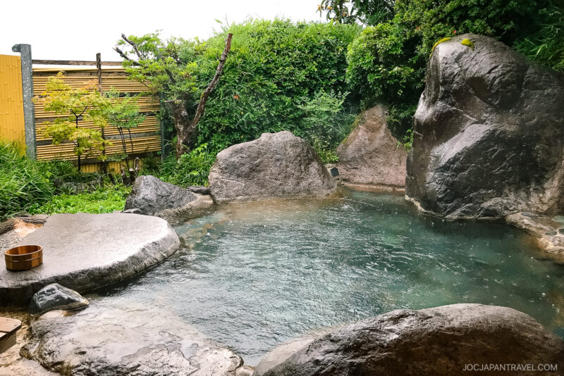 An outdoor Japanese onsen at Musouen Hotel, surrounded by large rocks, green bushes, and a bamboo fence. Steam rises gently from the clear hot spring water, with a wooden bucket resting at the pools edge.