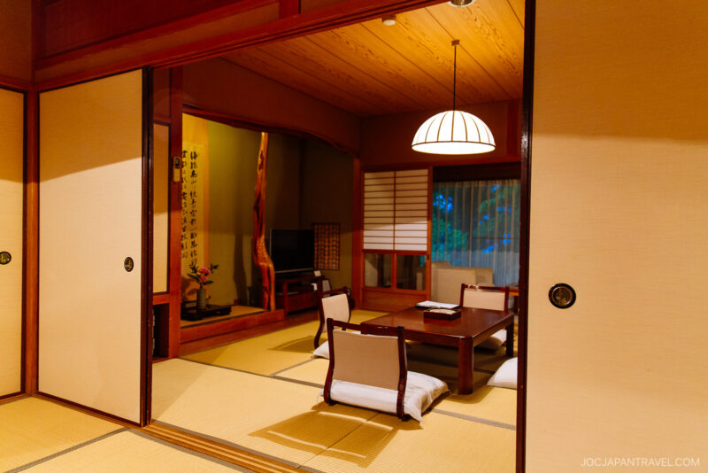 A traditional Japanese tatami room at Musouen Hotel with a low wooden table, floor chairs, sliding shoji doors, and soft lighting. A TV, scroll, and flower arrangement are visible in the background.