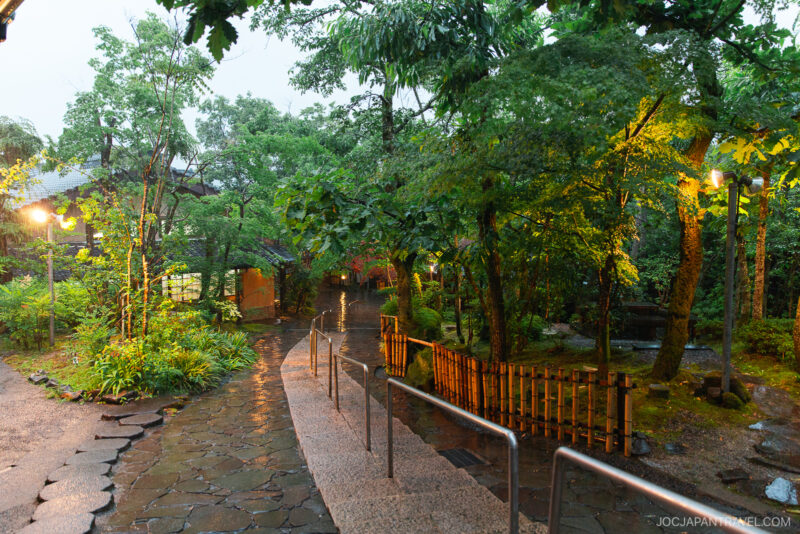 A gently curving stone path bordered by greenery and wooden fences leads through the lush, rain-soaked garden of Musouen Hotel, with glowing lamps illuminating the wet ground and trees.