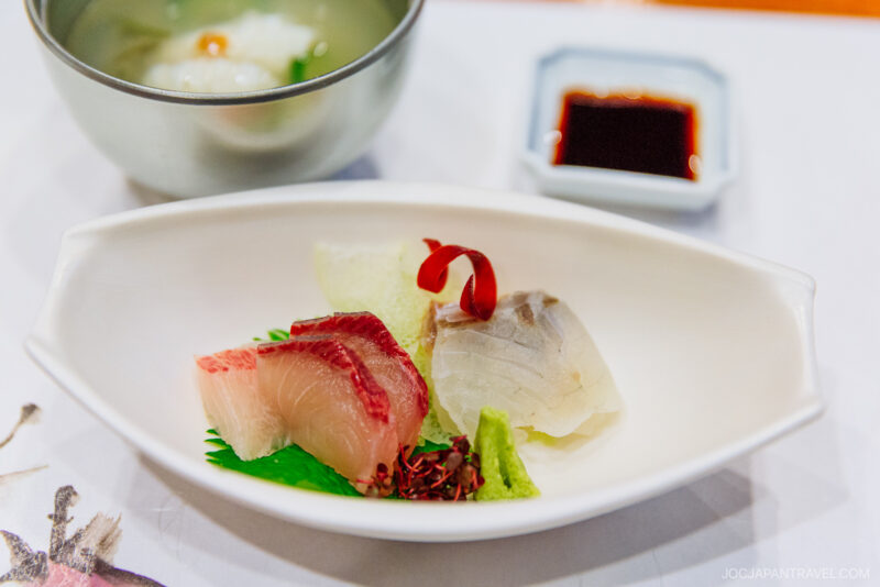 A white dish at Musouen Hotel features assorted sashimi slices, including tuna and white fish, garnished with wasabi and a red pepper strip. In the background are a small bowl of soup and a square dish of soy sauce.