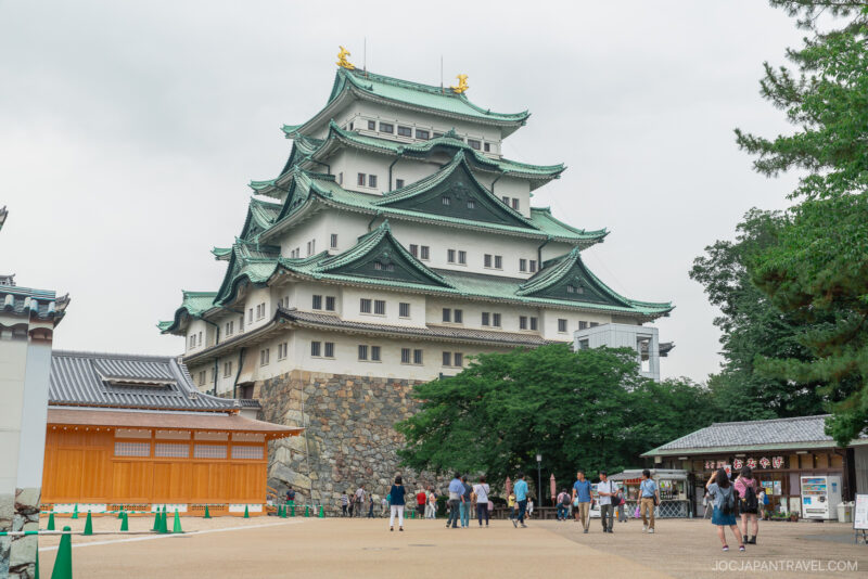 Nagoya Castle in Japan, with its iconic green-tiled roofs and golden ornaments, stands majestically as visitors stroll through the courtyard on a cloudy day.