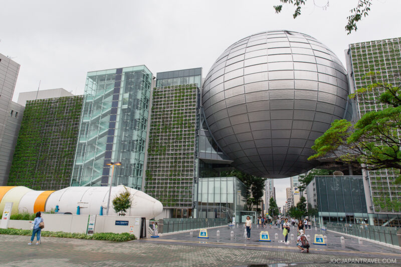A modern science museum, the Nagoya City Science Museum features a large silver sphere suspended between glass buildings, lush green walls, and people walking in front on a cloudy day.