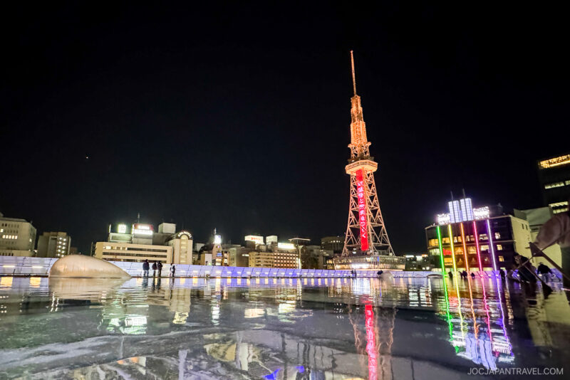 The Nagoya TV Tower glows in red and white against the night sky, its reflection shimmering on the wet ground. City lights and buildings create a vibrant backdrop as people stroll near the railing.