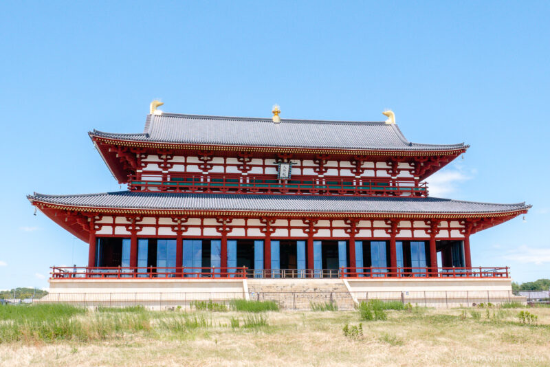 A large, traditional Japanese building at the Nara Palace Site with red columns, white walls, and a dark tiled roof stands against a clear blue sky, with grass in the foreground.