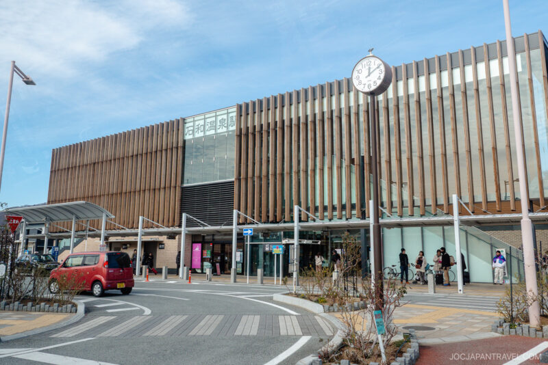 A modern train station building with vertical wooden slats at Isawa Onsen Yamanashi, a large clock in front showing 12:24, parked cars, bicycles, and people walking near the entrance under a blue sky.