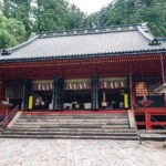 Traditional Japanese temple with a sloped roof, red pillars, stone steps, and two stone lanterns at the entrance, surrounded by lush green trees—evoking the serene beauty of Nikko Futarasan Jinja.