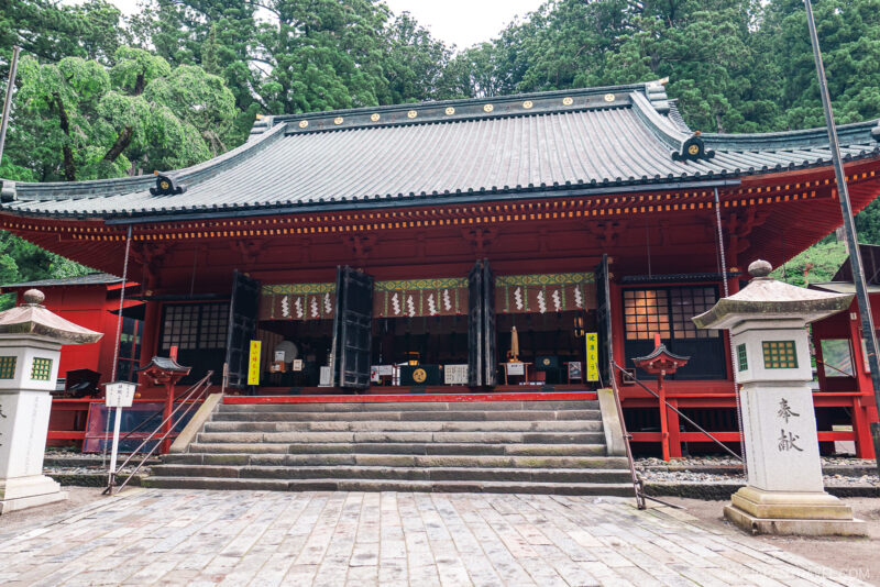 Traditional Japanese temple with a sloped roof, red pillars, stone steps, and two stone lanterns at the entrance, surrounded by lush green trees—evoking the serene beauty of Nikko Futarasan Jinja.