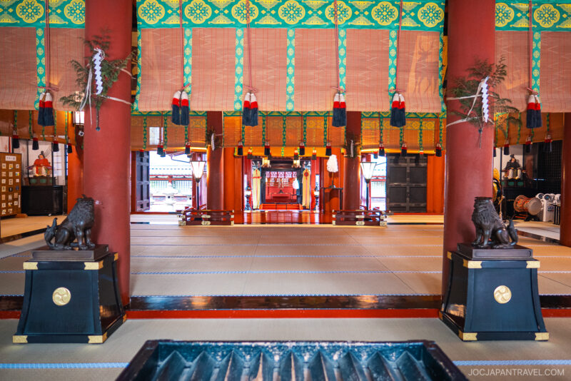 Interior of Nikko Futarasan Jinja, showcasing tatami mats, red pillars, decorative banners, and lion statues on pedestals. The altar area is visible in the background beneath ornate, colorful ceiling panels.